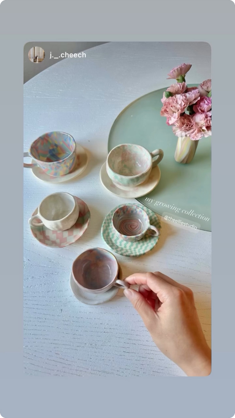 A collection of cute pastel themed nerikomi coffee mug and saucer set on top of table with a bud vase with pink flowers