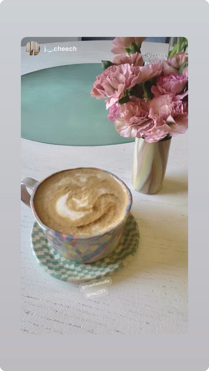 rainbow pastel multi color coffee mug on top of a pastel green checker saucer, nerikomi bud vase with pink flowers on top of a dining table